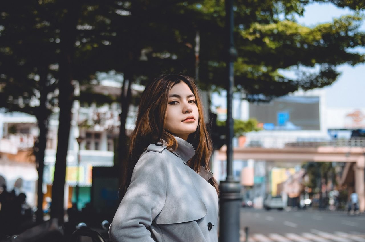 Home Young woman in a coat posing confidently in an urban street surrounded by trees.